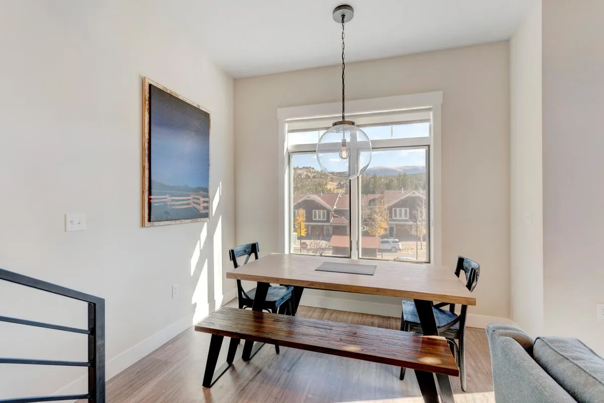 A warm and inviting dining area featuring a sturdy table set for gatherings, illuminated by a stylish hanging light fixture. Large windows let in natural light, while tasteful artwork adds a touch of personality. The space is clearly designed for social meals, games, or group conversations.