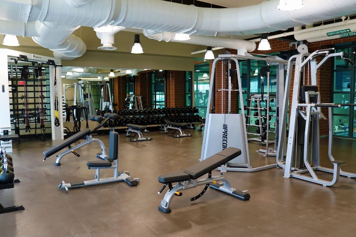 A well-equipped fitness room with weight lifting equipment, benches, and mirrored walls, designed for both individual and group workouts. The mood is motivational, suggesting opportunities for personal fitness and community.
