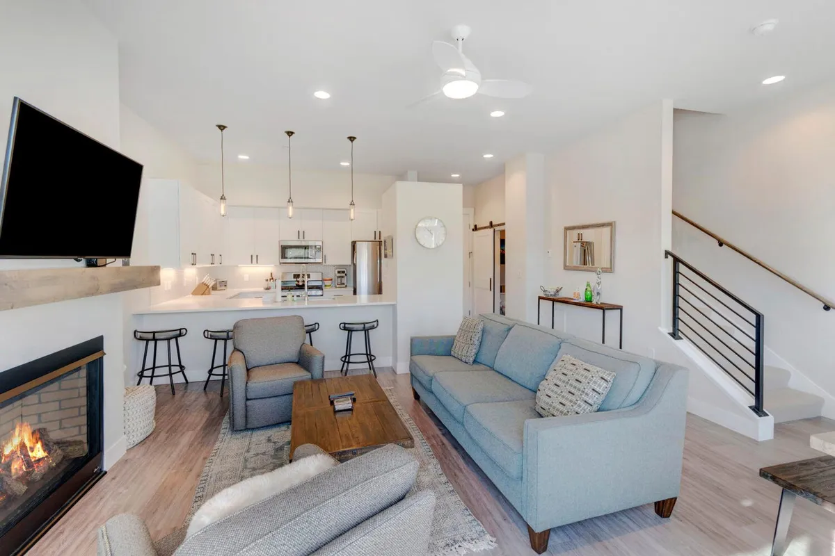The image showcases a stylish, open-concept living area featuring a modern fireplace, a plush sofa set perfect for gathering, a sleek kitchen island with elegant hanging lights, all set within cozy, contemporary decor. The space radiates warmth and comfort, inviting guests to relax by the fire, entertain friends, or enjoy meals together.