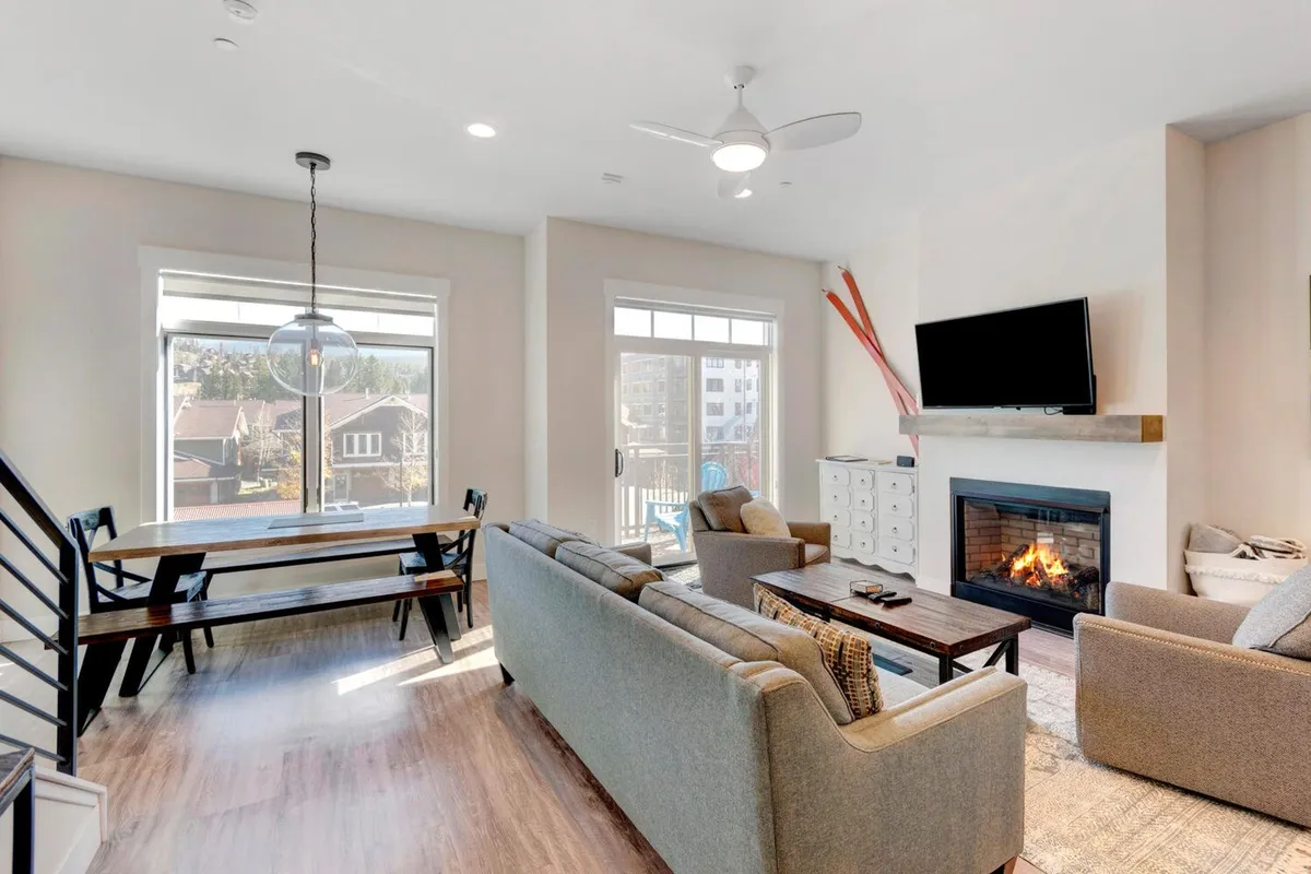 A wide shot of a bright, open interior that highlights a modern fireplace, large windows, a comfortable sofa, and a spacious dining table. Sunlight pours in, creating an inviting space ideal for family gatherings, relaxation, and entertaining by the fire. The arrangement naturally draws people together, echoing the sense of connection and laughter described.