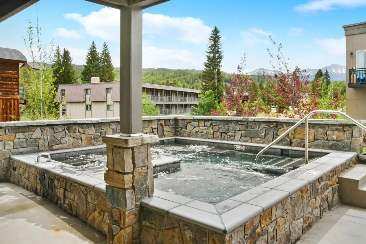 A spacious, stone-surrounded hot tub set amidst greenery, with people relaxing and appreciating the tranquil atmosphere. The setting offers a resort-style retreat after a day of adventure.