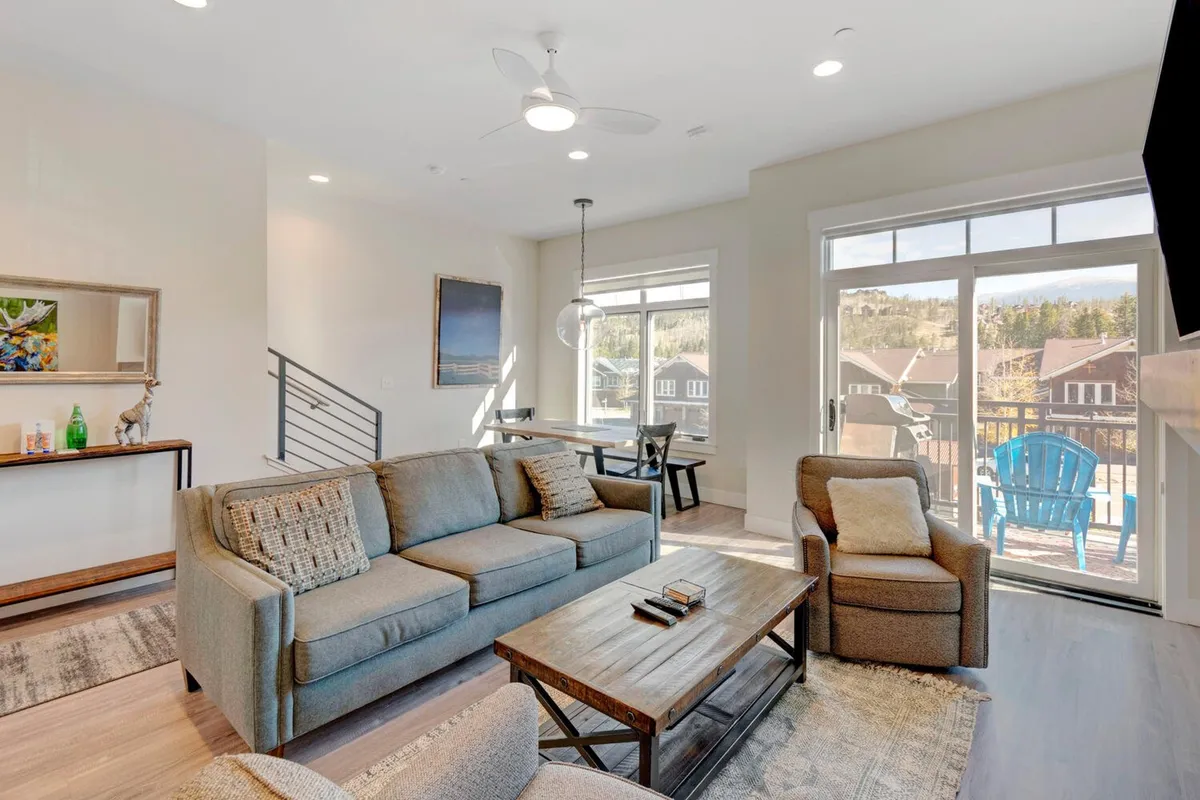 This cozy living area is anchored by a plush sofa and blue accent chair, with a coffee table and large windows providing abundant natural light. The adjacent dining area is visible, blending seamlessly into the living lounge. The setting evokes movie nights, family bonding, and gatherings around the table after a day of adventure.