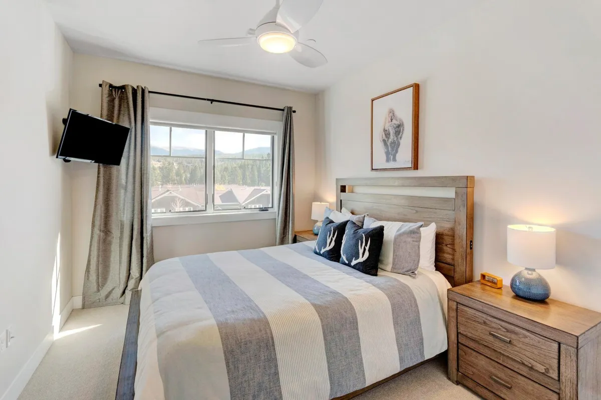 A serene bedroom featuring a comfortable bed with striped bedding, a large window allowing natural light and offering a picturesque mountain view, a nightstand with a lamp, and an art piece depicting a bear. The overall atmosphere is calm and inviting, ideal for restful nights and peaceful mornings.