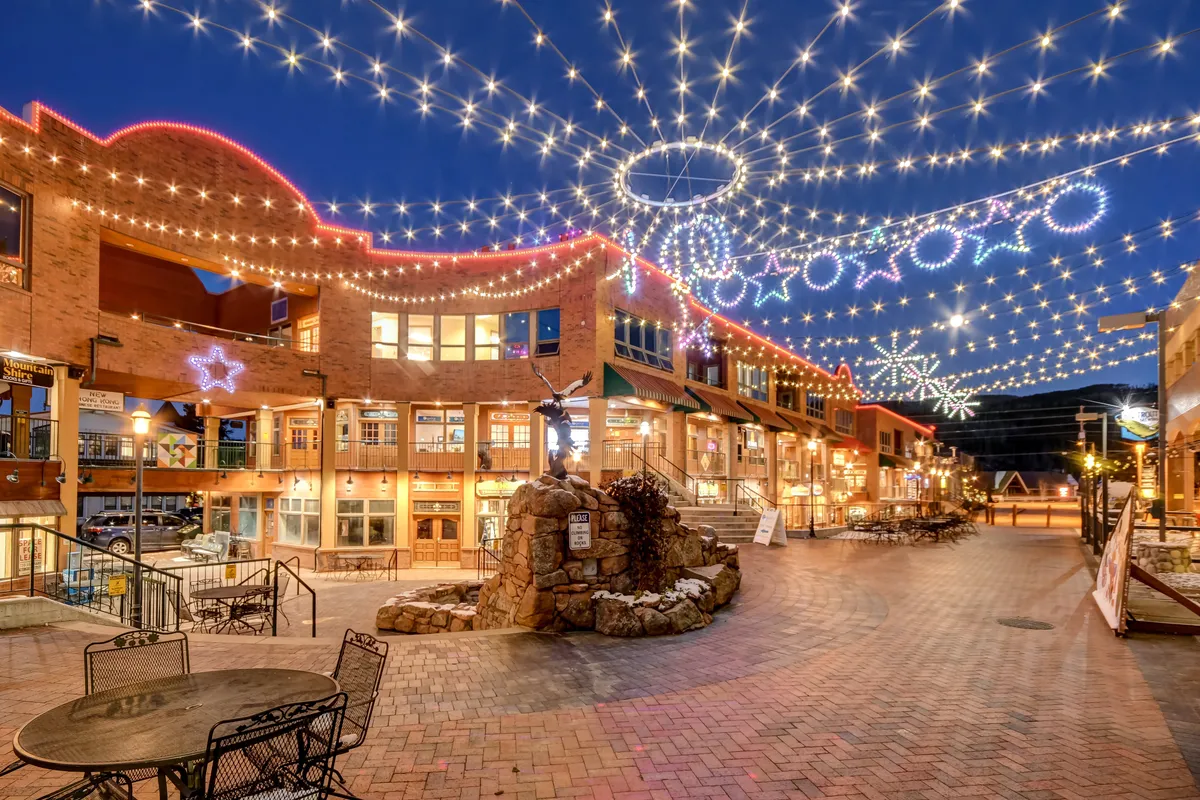 The image displays a vibrant outdoor plaza with string lights overhead, a stone fountain as a centerpiece, decorated buildings, and an inviting outdoor seating area. People are seen dining, shopping, and socializing, creating an atmosphere of celebration and community. The festive lights and lively environment suggest a curated and memorable local experience.