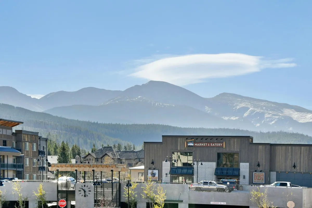 A scenic view of a mountain market area with a clock tower, outdoor dining tables, and trees. The mountains rise in the background, suggesting easy access to both natural and social experiences in all seasons.